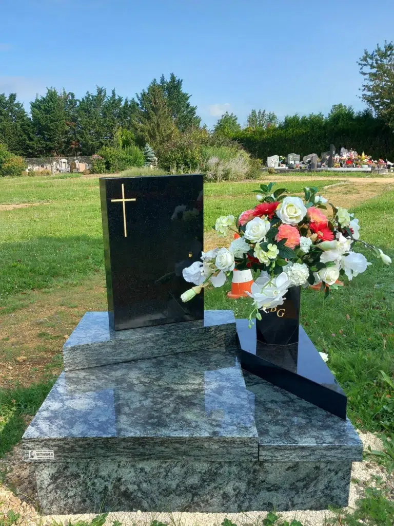 Monument funéraire en granite noir et gris avec croix dorée et fleurs.
